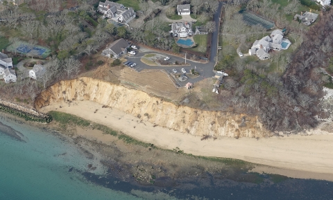an aerial view of an eroding coastal bluff at a national wildlife refuge. A parking lot and residential buildings are seen in the background