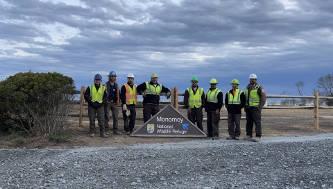 a group of maintenance staff in uniform stand in a gravel lot posing with a triangular sign for the Monomoy National Wildlife Refuge Headquarters