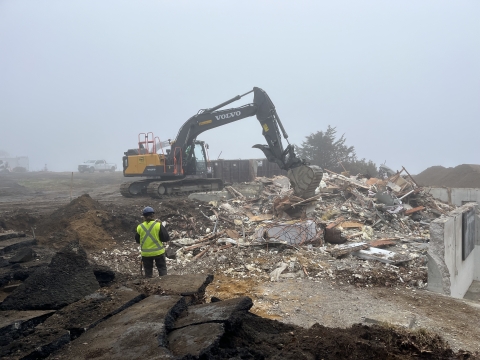A construction worker overlooks a tractor working on the demolished ruins of a building. The weather is foggy with low visibility