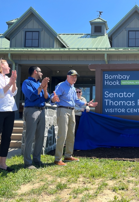 a group of people stand at a large visitor center facility. One of the people pulls off a blue fabric to unveil a sign that says "Bombay Hook Senator Thomas R. Carper Visitor Center". People are clapping in celebration of the event.
