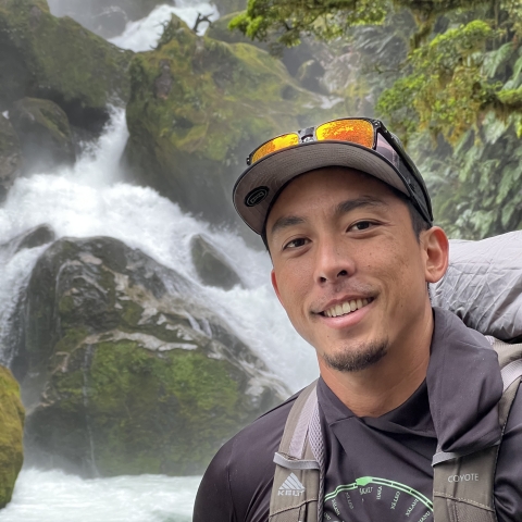 Koa stands in front of a waterfall. He is dressed in black with a black hat and sunglasses on the brim. He is holding a camera.