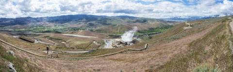 Wide view of a geothermal power plant nestled in the hills under a cloud covered sky with snow capped mountains in distance. 