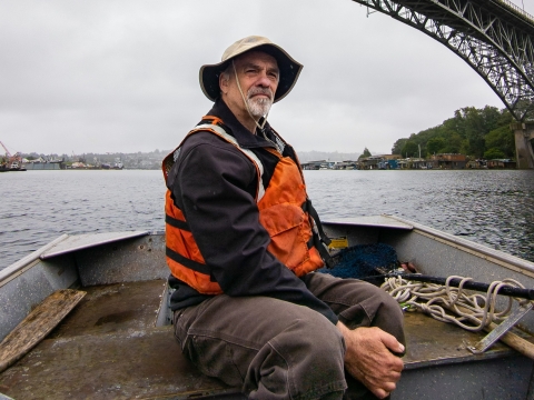 A man sits in a boat on water. He wears a brimmed hat and orange life vest. A bridge spans above him.