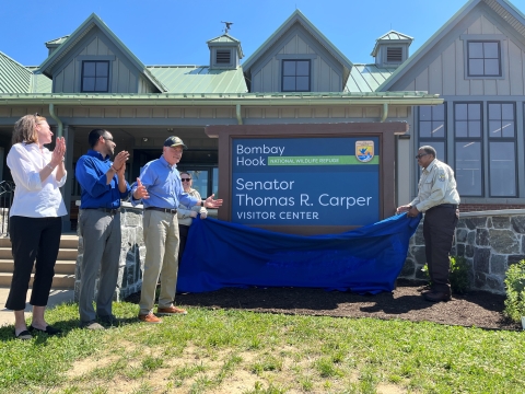 a group of people stand by while a sign is unveiled. The sign reads "Senator Thomas R. Carper Visitor Center". The people applaud while the Senator stands in acknowledgment
