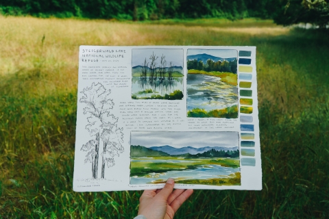 Artist hand (Claire Giordano) holding up their watercolor artwork in front of a scene at Steigerwald Lake NWR