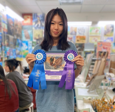 Young person in art studio holds winning duck stamp art adorned with two ribbons.