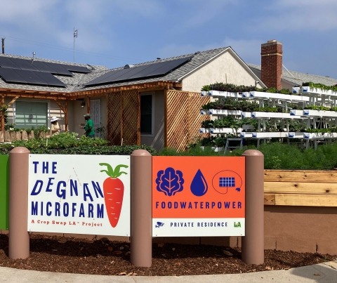 A house with solar panels on the roof and an extensive microfarm in the front yard. Signs read: The Degan Microfarm: A Crop Swap LA Project. Food, Water, Power, Private Residence.