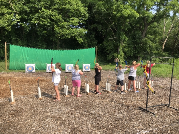 Six kids holding a bow and arrow. They are aiming at a target with a green backdrop. 