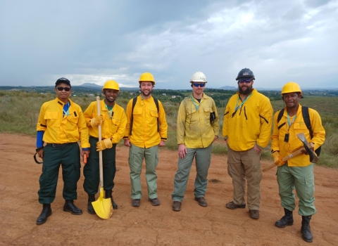 Six firefighters stand in a line for a photo. They are all in yellow nomex and fire pants. They are standing on a dry road with dry vegetation behind them.