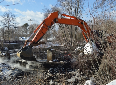 The orange arm of a construction excavator reaches into a river to break apart a dam
