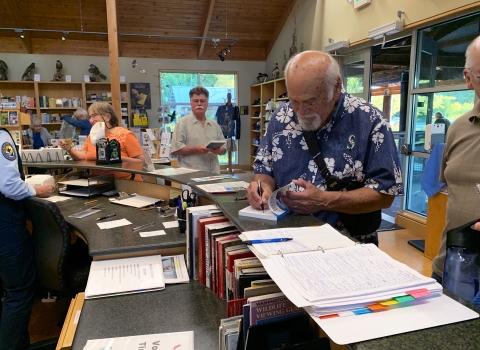 A man with white hair and beard and wearing a Hawai'ian shirt signs a book at the counter of a visitor center store.